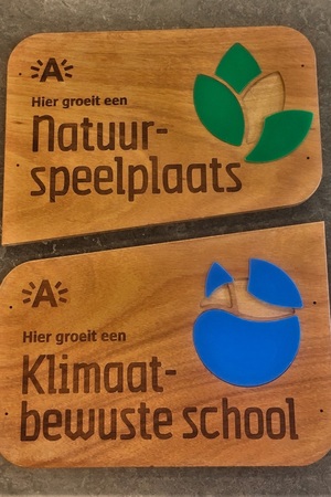 Twee schoolpoortborden uit hout. Op de ene staat "hier groeit een natuurspeelplaats" en op de andere "hier groeit een klimaatbewuste school". Twee schoolpoortborden uit hout. Op de ene staat "hier groeit een natuurspeelplaats" en op de andere "hier groeit een klimaatbewuste school".