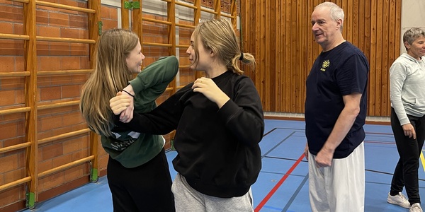 Twee meisjes leren technieken om zichzelf te verdedigen. Een leraar kijkt toe.