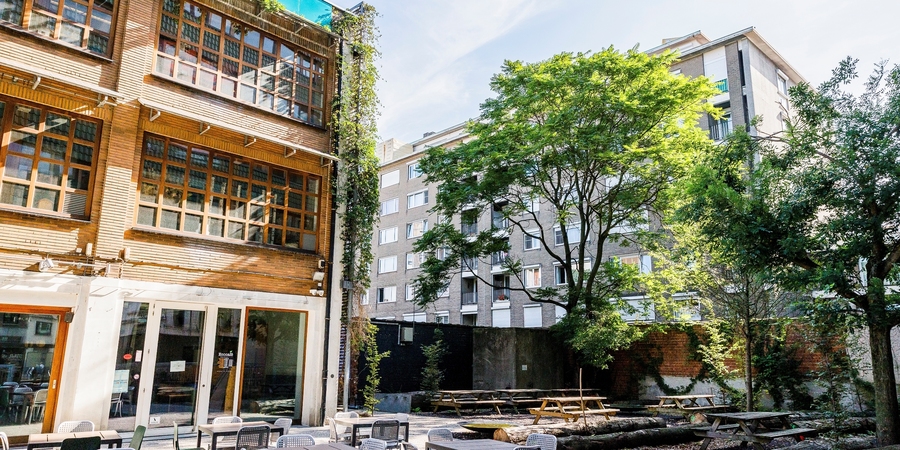 We zien het voorplein van het EcoHuis, met het terras en picknicktafels en veel groen.