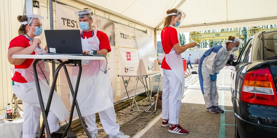 Twee medewerkers van testdorp TestCovid staan aan een tafeltje waarop een laptop staat. Twee anderen nemen een test af van iemand die in een auto zit. Twee medewerkers van testdorp TestCovid staan aan een tafeltje waarop een laptop staat. Twee anderen nemen een test af van iemand die in een auto zit.