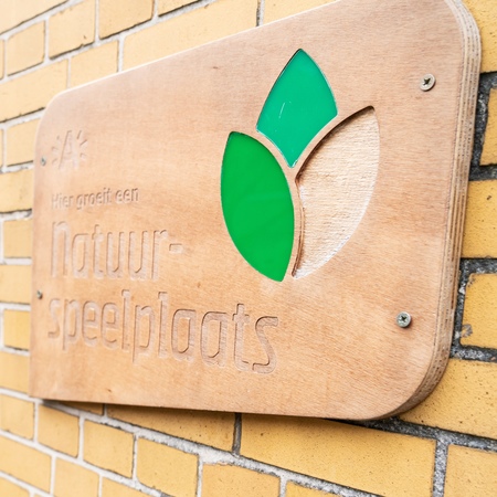Een houten schoolpoortbord aan een schoolgevel. Op het bord staat "hier groeit een natuurspeelplaats". Een houten schoolpoortbord aan een schoolgevel. Op het bord staat "hier groeit een natuurspeelplaats".