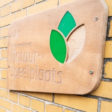Een houten schoolpoortbord aan een schoolgevel. Op het bord staat "hier groeit een natuurspeelplaats". Een houten schoolpoortbord aan een schoolgevel. Op het bord staat "hier groeit een natuurspeelplaats".