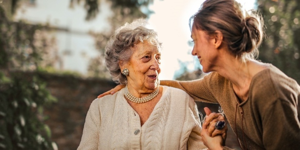 Een vrouwelijke mantelzorger legt haar arm om een bejaarde vrouw terwijl ze beiden glimlachen.