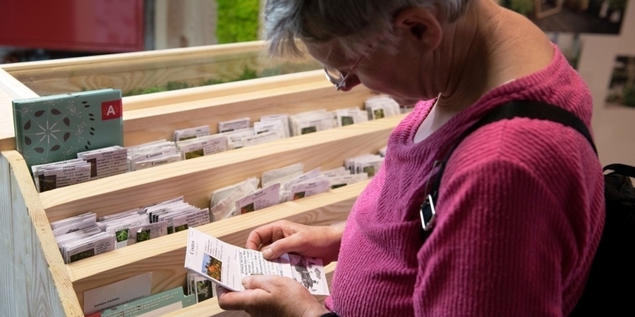 Een vrouw bekijkt een pakje zaadjes in de zadenbib en leest de bijbehorende uitleg. Een vrouw bekijkt een pakje zaadjes in de zadenbib en leest de bijbehorende uitleg.