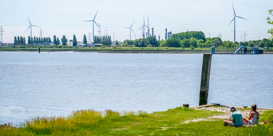 Een man en een vrouw zitten op een stukje groen langs de Schelde. Aan de overkant staan enkele windmolens. Een man en een vrouw zitten op een stukje groen langs de Schelde. Aan de overkant staan enkele windmolens.
