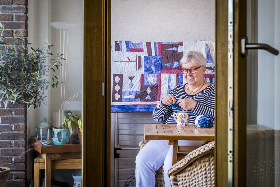 Vrouw zit op terras te haken. We zien duidelijk de dikke houten frames van de driedubbele beglazing. Vrouw zit op terras te haken. We zien duidelijk de dikke houten frames van de driedubbele beglazing.