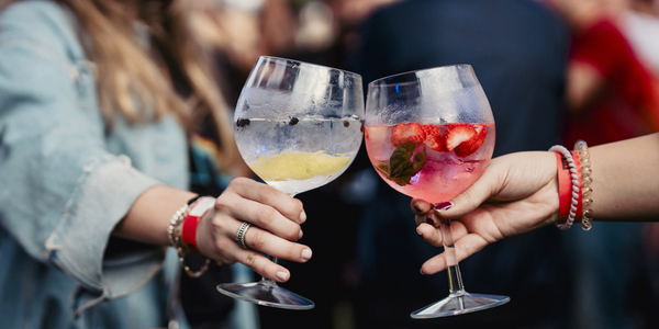 Twee mensen proosten met glazen gevuld met kleurrijke drankjes, een met citroen en ijs, de ander met aardbeien en munt. De achtergrond is onscherp, wat de focus op de glazen legt.