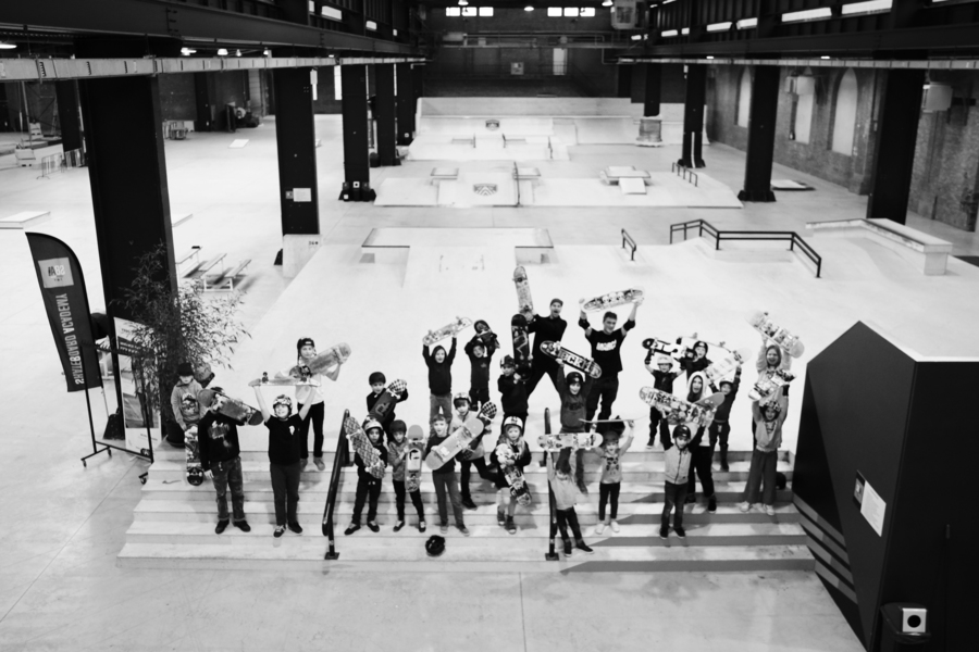 Zwart wit foto van een groep jonge skaters die voor het skatepark staan en hun skateboard in de lucht steken.