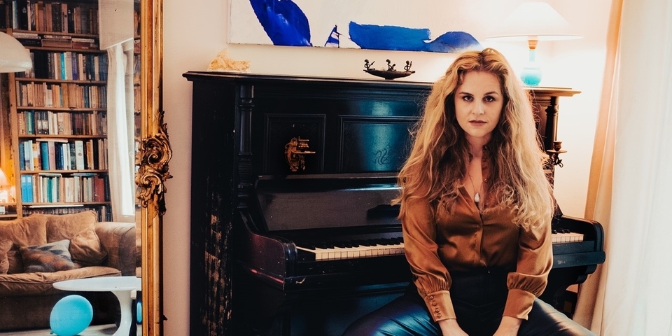 Fleur Van Groningen op een pianokruk voor een piano, een spiegel reflekteerd een bruine zetel Fleur Van Groningen op een pianokruk voor een piano, een spiegel reflekteerd een bruine zetel