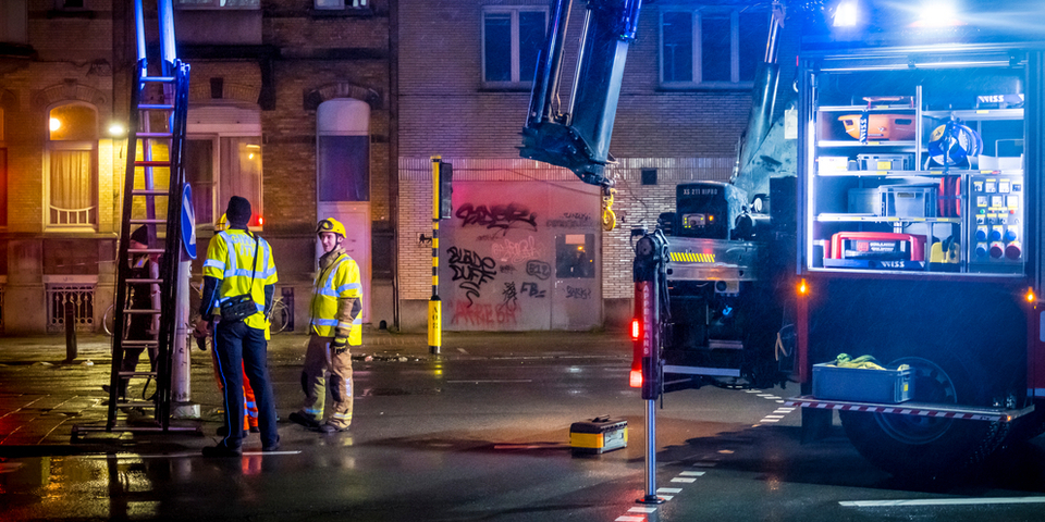 Twee brandweermannen staan met een brandweerladder voor enkele woningen in een straat.