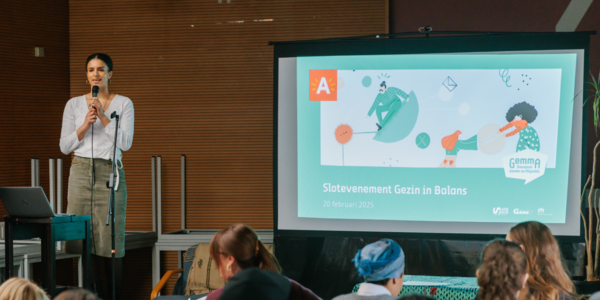 Projectleider Marie Kenis houdt een microfoon vast, naast haar wordt een presentatie geprojecteerd