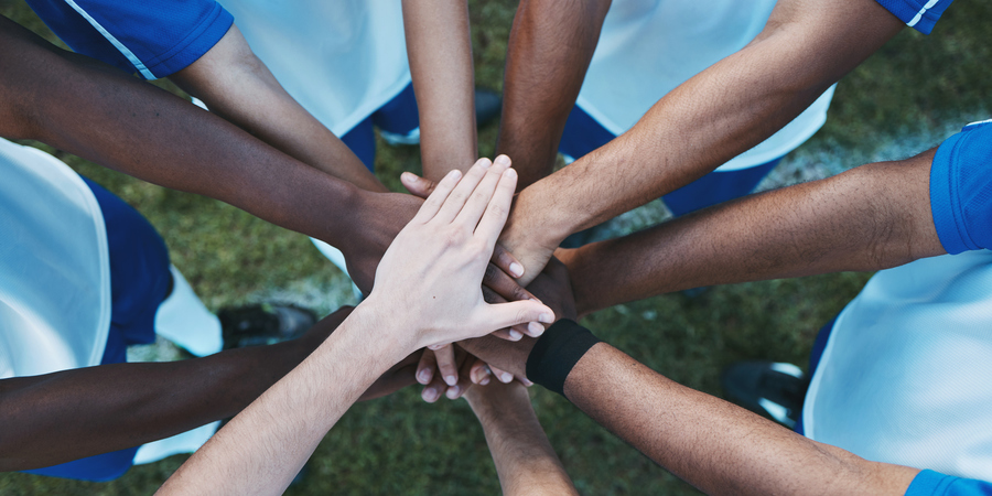 Een groep mensen in sportkleding staat in een cirkel en legt hun handen op elkaar – een krachtig symbool van teamgeest, samenwerking en gedeelde motivatie.