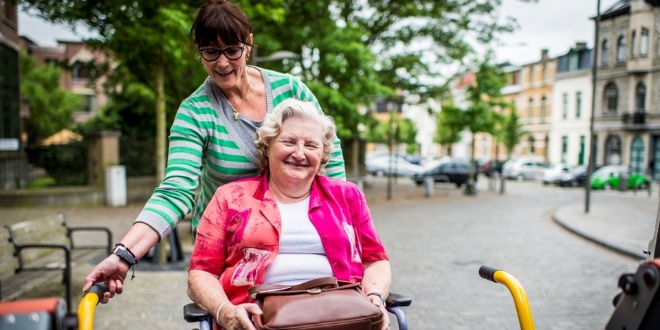 Een dame helpt een oudere dame in haar rolstoel in te stappen in een busje voor mindervaliden.