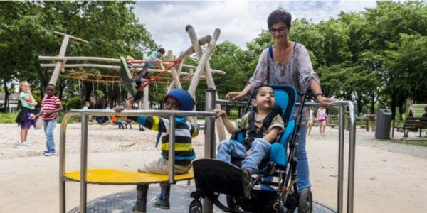 Speeltuin in een park met spelende kinderen. Een jongen met een beperking in een rolstoel speelt mee en wordt begeleid door een volwassen vrouw.