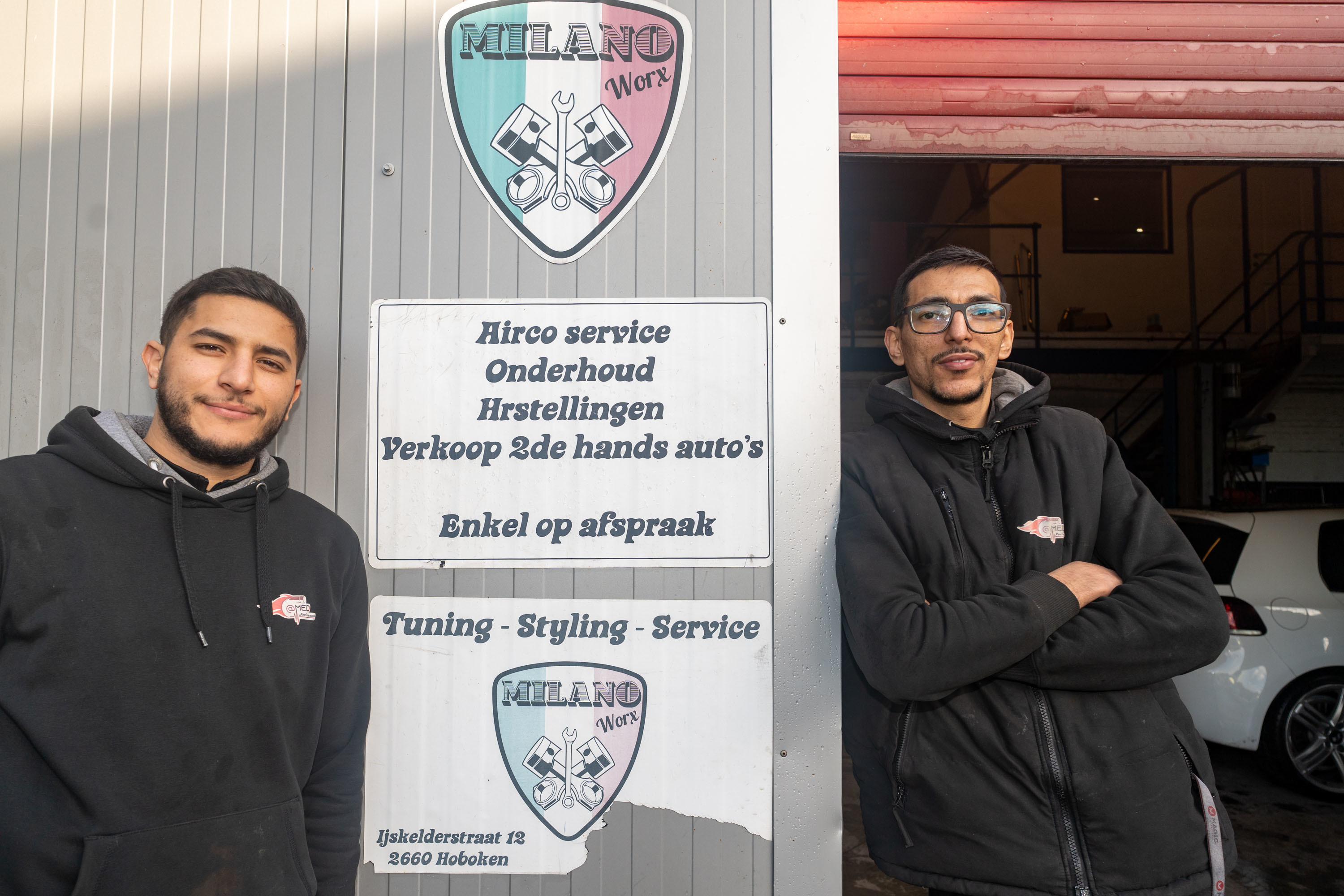 Het duo poseert aan de ingang van de garage bij de identiteitsplaat met vermelding 'Onderhoud en herstelling en Milano Worx'' Het duo poseert aan de ingang van de garage bij de identiteitsplaat met vermelding 'Onderhoud en herstelling en Milano Worx''