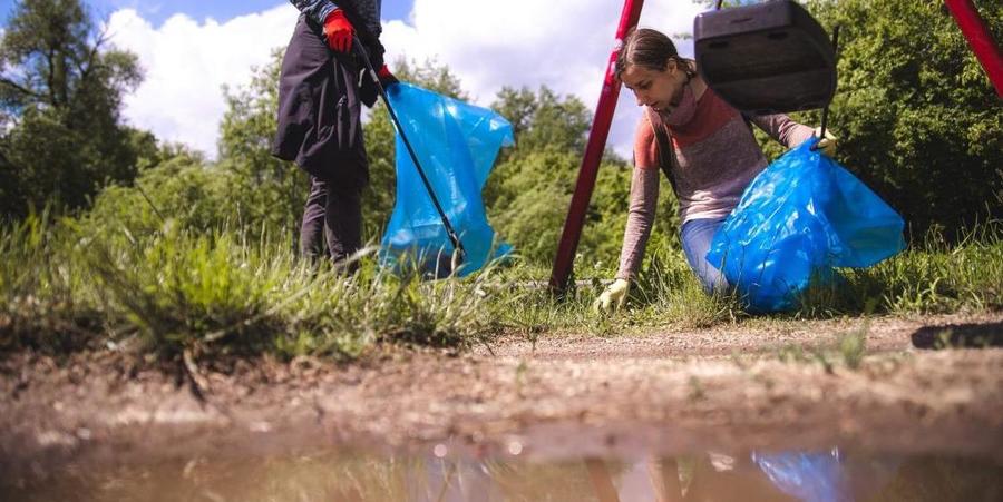 Twee mensen met blauwe vuilniszakken ruimen zwerfvuil op langs een rivier.