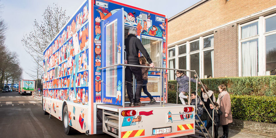 Kinderen en volwassenen stappen in de bibmobiel, een kleine vrachtwagen die is omgevormd tot rijdende bibliotheek. Kinderen en volwassenen stappen in de bibmobiel, een kleine vrachtwagen die is omgevormd tot rijdende bibliotheek.