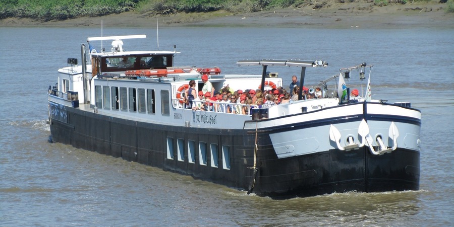 Tientallen mensen zitten op het dek van een binnenschip dat aan het varen is. Tientallen mensen zitten op het dek van een binnenschip dat aan het varen is.