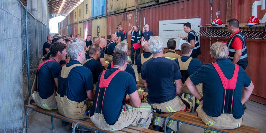 Veel brandweermannen op banken die naar whiteboard kijken met een lesgever