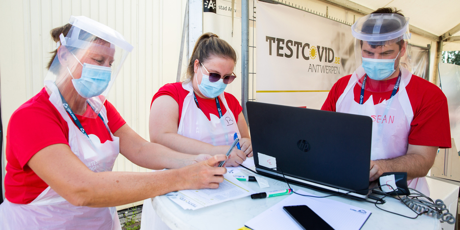 Drie medewerkers van het testdorp TestCovid staan aan een tafel waarop een laptop staat en enkele papieren liggen. Drie medewerkers van het testdorp TestCovid staan aan een tafel waarop een laptop staat en enkele papieren liggen.