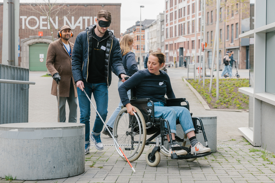 een geblinddoekte man met een wandelstok houdt een rolstoel vast waar een vrouw in zit, een man met koptelefoon loopt daar achter