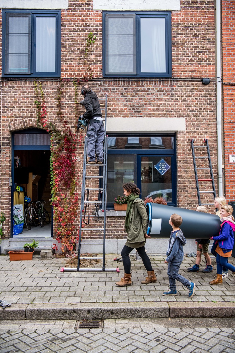 Een gezin draagt samen een regenton naar hun huis. Op de achtergrond wordt een klimhulp geïnstalleerd.