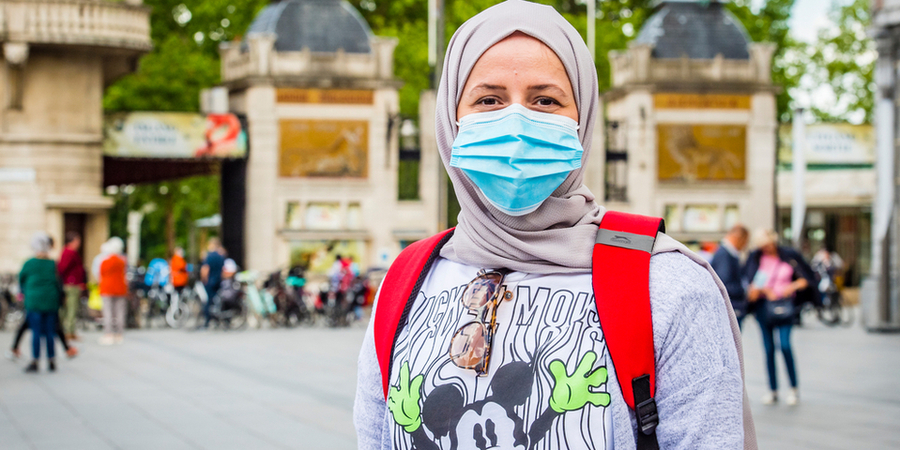 Een vrouw met mondmasker die voor de ZOO staat, kijkt in de camera. Op de achtergrond staan tientallen mensen aan de ingang. Een vrouw met mondmasker die voor de ZOO staat, kijkt in de camera. Op de achtergrond staan tientallen mensen aan de ingang.