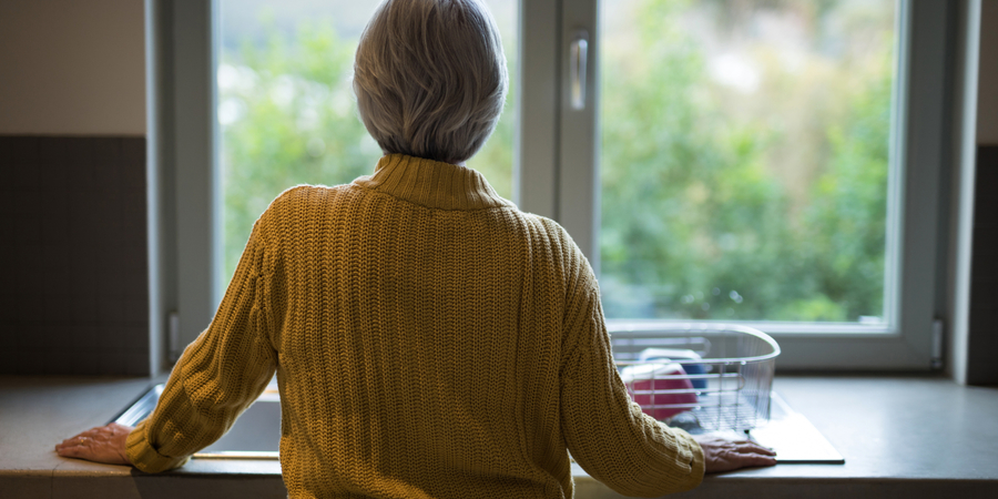 Een vrouw met grijs haar en een gele trui staat met haar rug naar de camera. Ze kijkt door het raam naar buiten. Een vrouw met grijs haar en een gele trui staat met haar rug naar de camera. Ze kijkt door het raam naar buiten.