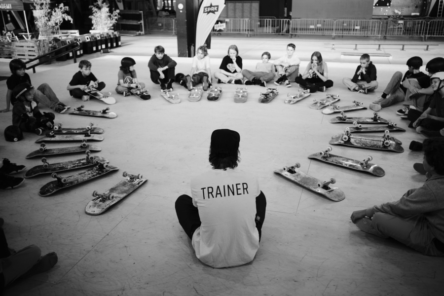 Een trainer en een 15-tal jongeren zitten in een cirkel. Hun skateboad ligt voor hen.