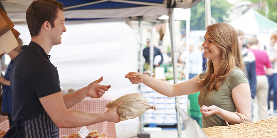 Een persoon koopt gebak bij een kraam op een drukke markt. De verkoper draagt een schort en de koper heeft een rieten tas. Op de achtergrond zijn meer kramen en mensen te zien.
