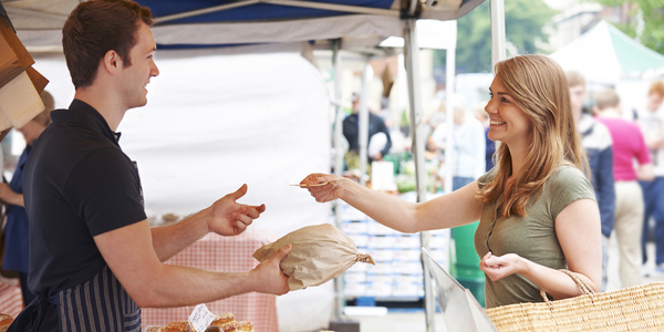 Een persoon koopt gebak bij een kraam op een drukke markt. De verkoper draagt een schort en de koper heeft een rieten tas. Op de achtergrond zijn meer kramen en mensen te zien.