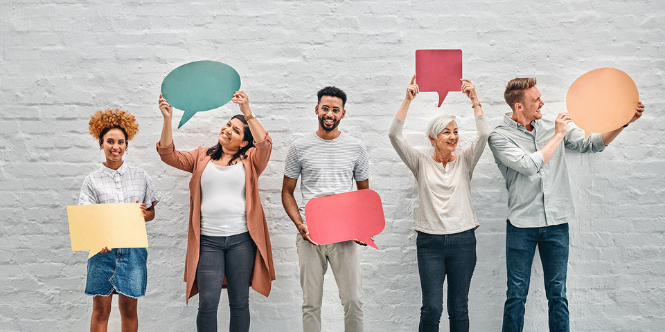 Vijf mensen staan voor een witte bakstenen muur, elk met een leeg spraakballonbord in een andere kleur: geel, groen, rood, rood en oranje.