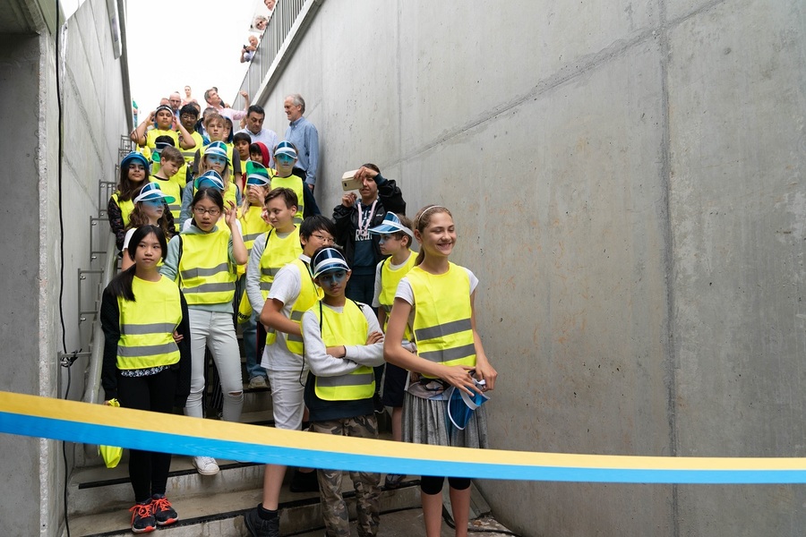 Schoolkinderen met fluo hesjes aan staan op de trap naar de tunnel achter een blauw-geel lint te wachten tot de tunnel geopend is.