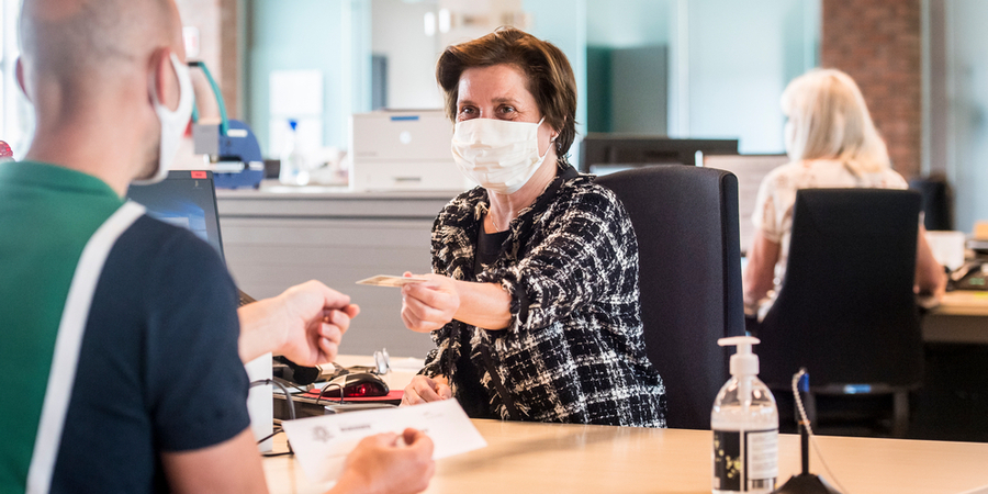 Een klant van een stadsloket geeft haar identiteitsbewijs aan een loketmedewerker. Beiden dragen een mondmasker. Een klant van een stadsloket geeft haar identiteitsbewijs aan een loketmedewerker. Beiden dragen een mondmasker.